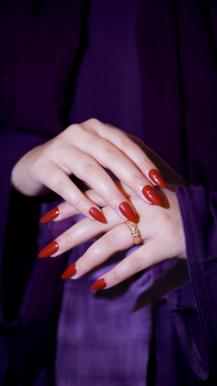 Matte deep-red almond press-on gel nails on both hands resting against a draped purple silk — Mona's signature work